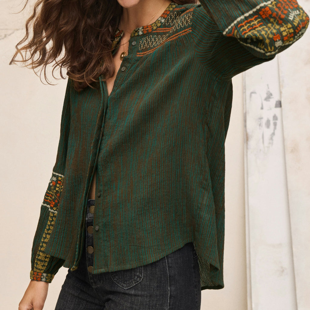 Woman wearing a green blouse with intricate patterns, standing against a neutral background.