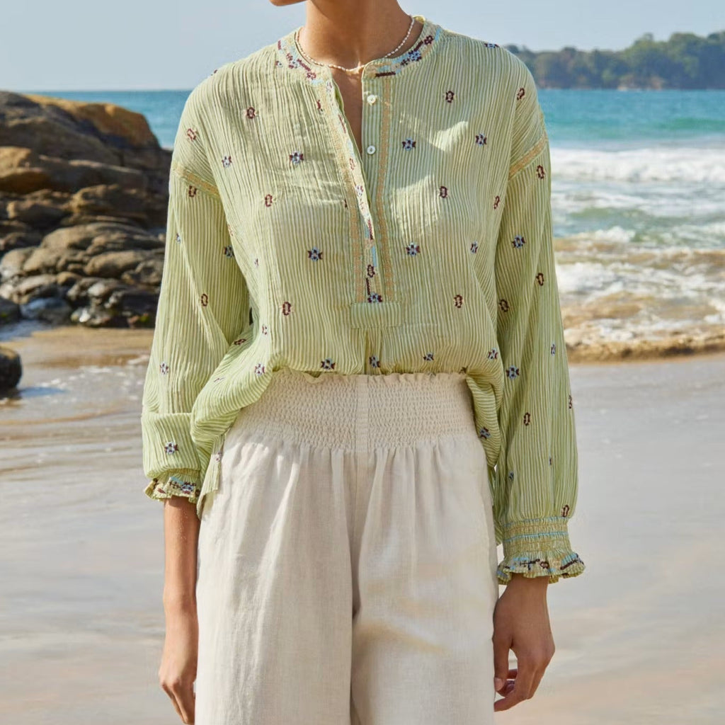 Woman in light green top and white pants standing on a beach with ocean and rocks in the background