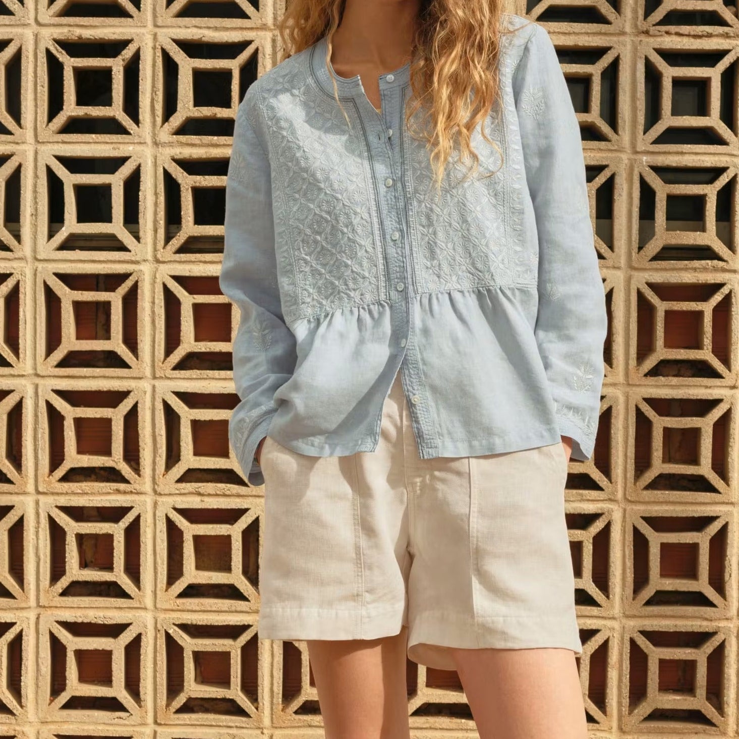 Woman wearing a light blue blouse and beige shorts standing against a geometric-patterned wall.