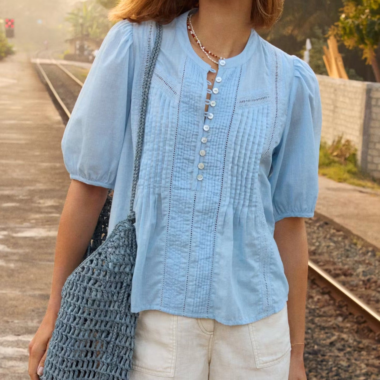 Woman standing on a train platform wearing a blue shirt and white shorts, holding a blue bag.